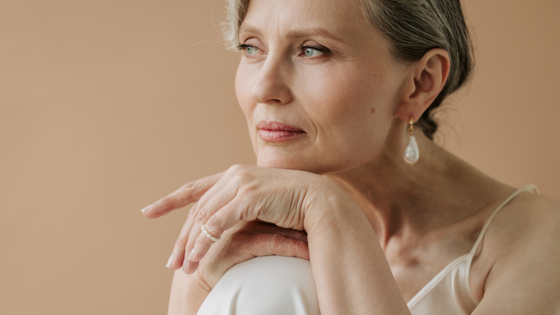 Woman in a cream top resting her chin on her hand against a warm beige backdrop
